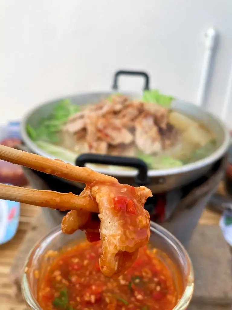 Hand dipping meat in Thai barbecue sauce with a pot of mookata in the distance.