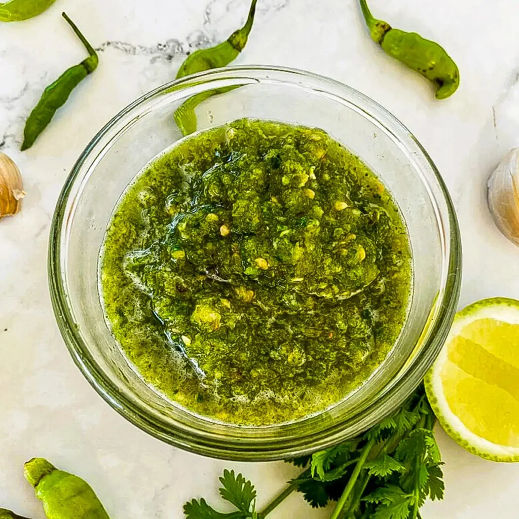 Close-up of Thai green chili sauce for seafood in a clear bowl, with green chilies, a lime wedge, and fresh herbs on a marble surface.