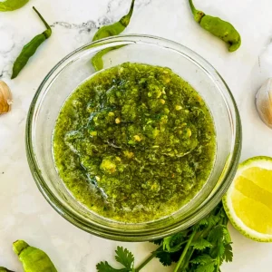 Close-up of Thai green chili sauce for seafood in a clear bowl, with green chilies, a lime wedge, and fresh herbs on a marble surface.