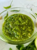 Close-up of Thai green chili sauce for seafood in a clear bowl, with green chilies, a lime wedge, and fresh herbs on a marble surface.