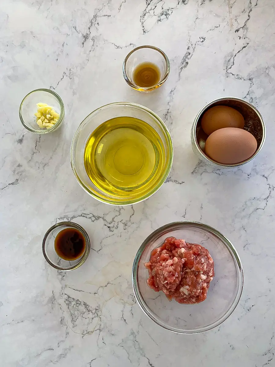 Ingredients for Thai pork omelette with eggs, ground pork, garlic, fish sauce, and oil laid out on a counter.