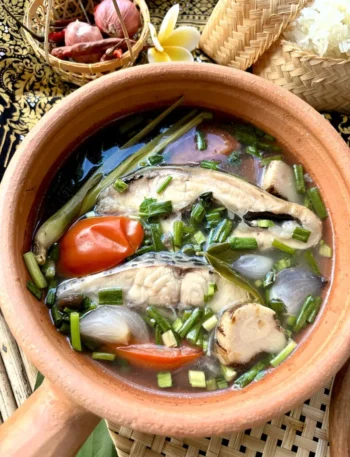 Tom yum pla, a Thai fish soup, with a rich broth, fish slices, and fresh herbs. Served in a clay pot with a side of Thai sticky rice.