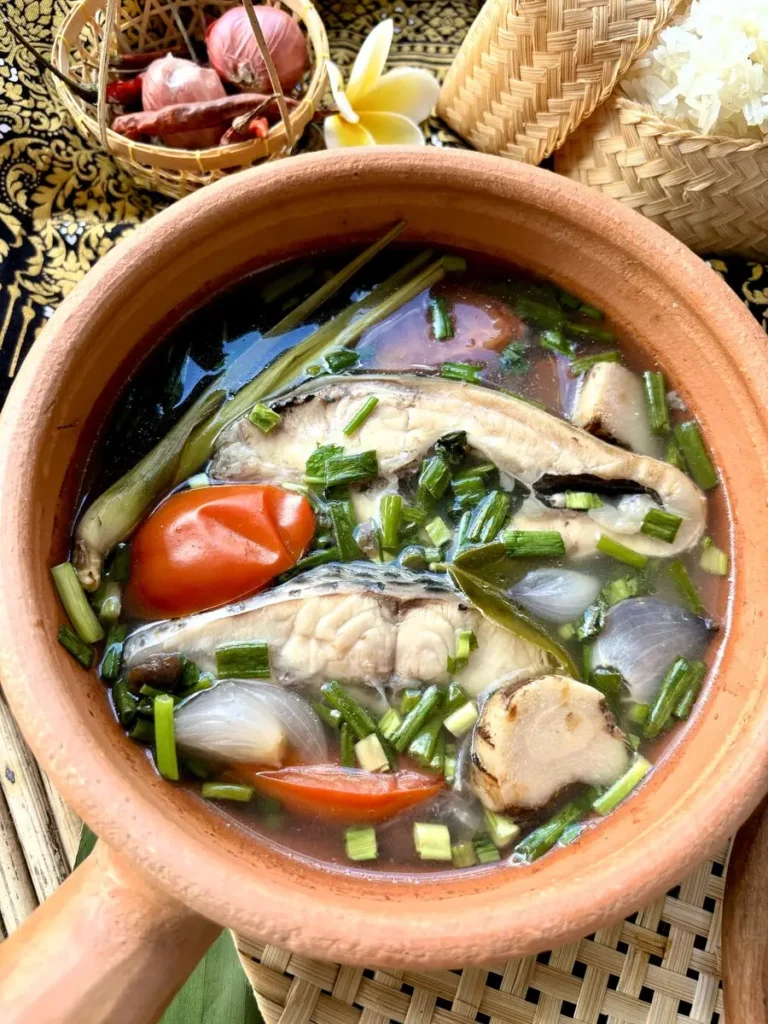 Tom yum pla, a Thai fish soup, with a rich broth, fish slices, and fresh herbs. Served in a clay pot with a side of Thai sticky rice.