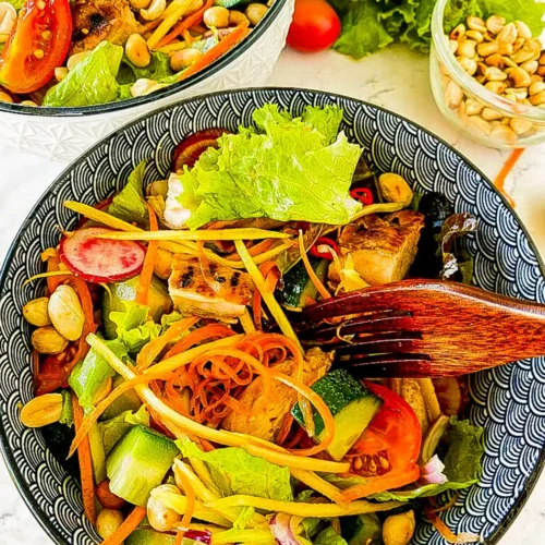 Spicy Thai chicken salad in a patterned black bowl with grilled chicken, lettuce, peanuts, cucumber, and shredded vegetables.