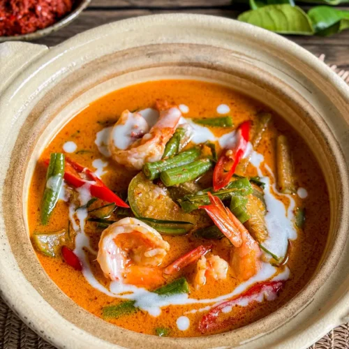 Thai red curry with shrimp and vegetables in coconut milk, topped with coconut cream, red chili, and long beans in a clay bowl.