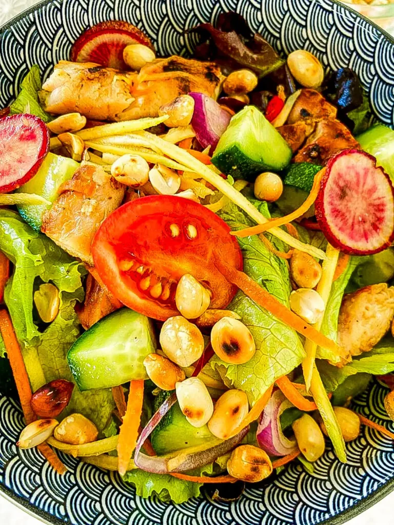 Close-up of Thai spicy chicken salad with sliced radish, cherry tomato, grilled chicken, peanuts, cucumber, and lettuce in a black bowl.