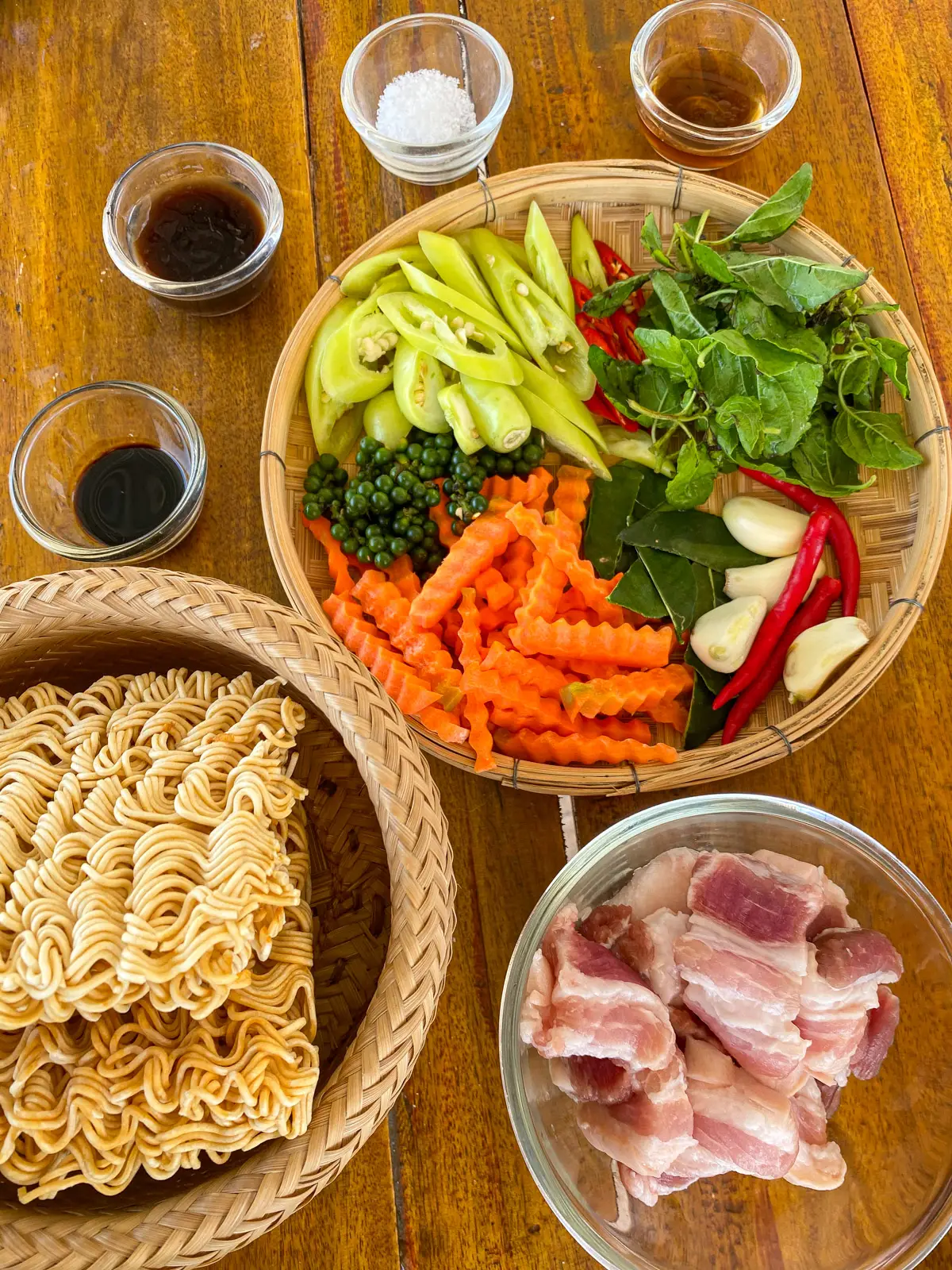 Ingredients for drunken Mama noodles, including Mama instant noodles, pork belly, holy basil, fresh chilies, peppercorns, kaffir lime leaves, carrots, and Thai stir-fry sauces.