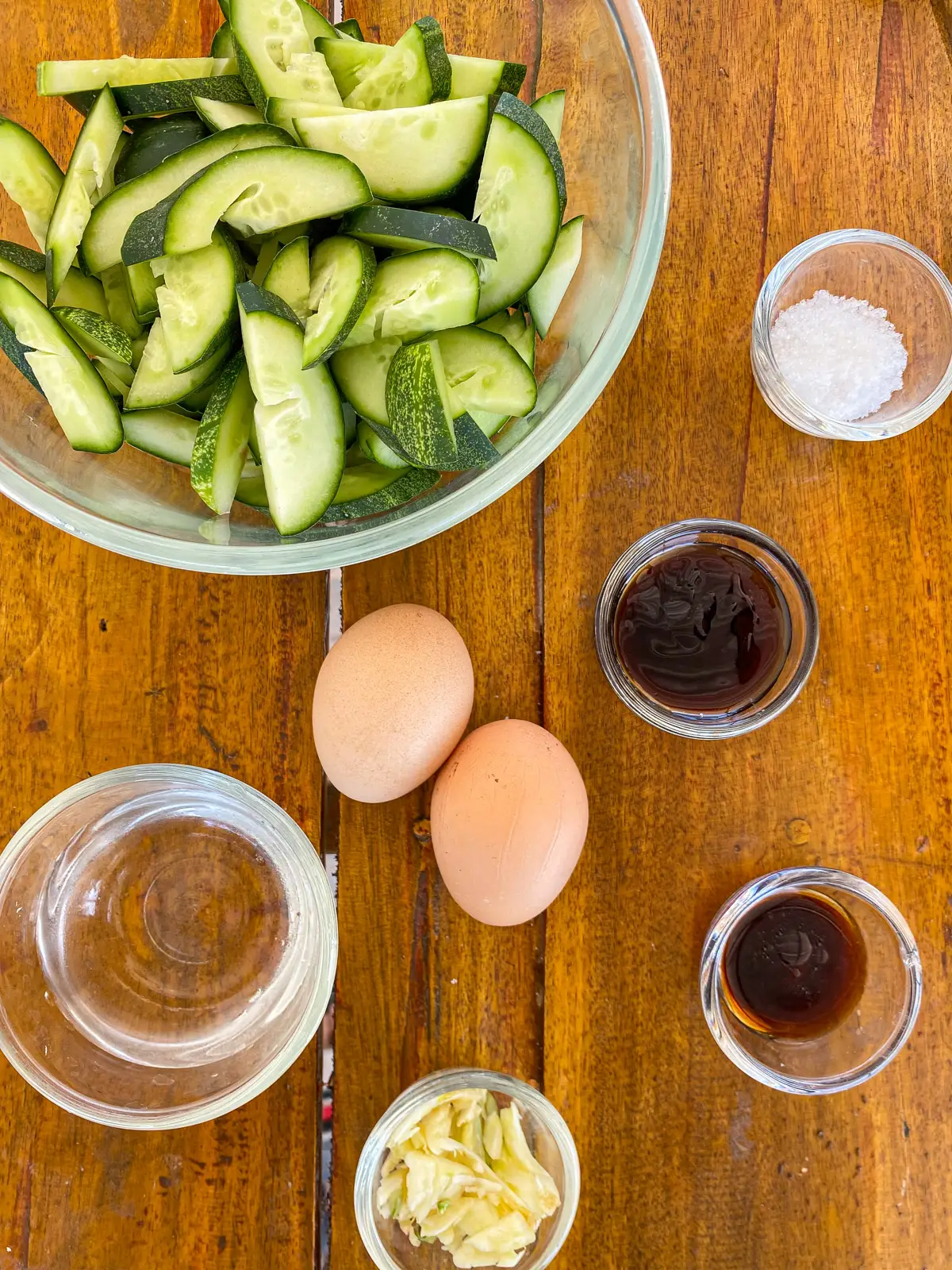 Top-view of ingredients for cucumber and egg stir-fry: cucumber slices, white sugar, oyster sauce, golden mountain sauce, eggs, garlic, and water.