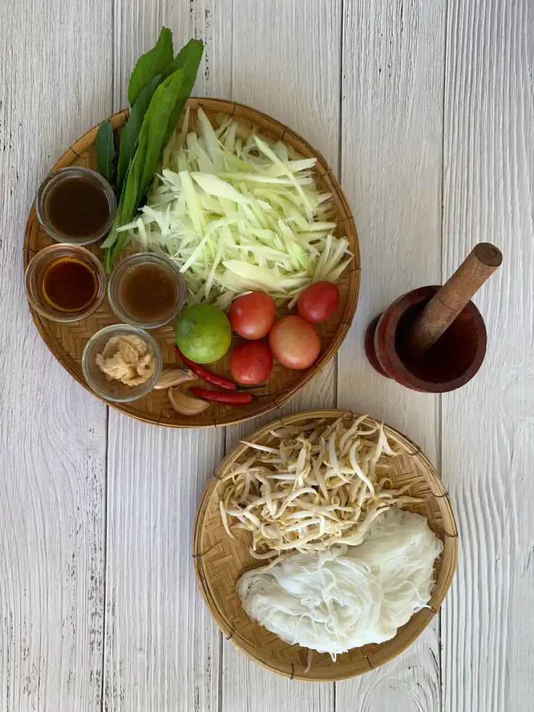 Ingredients for tam sua laid out on bamboo plates including shredded green papaya, rice vermicelli, tomatoes, and condiments with a mortar and pestle.