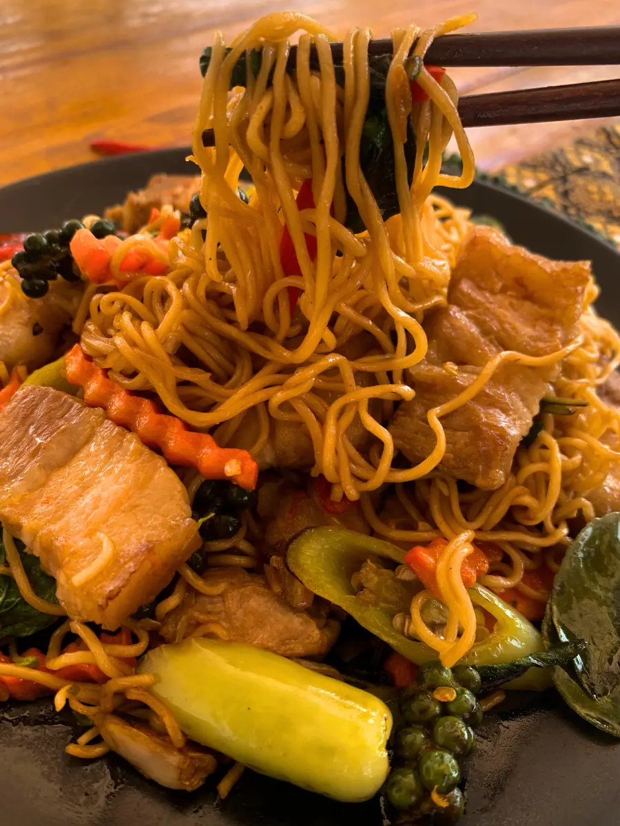 Chopsticks lifting a heap of pad kee mao instant noodles stir-fried with chunks of bacon, crisp vegetables, and Thai chilies.