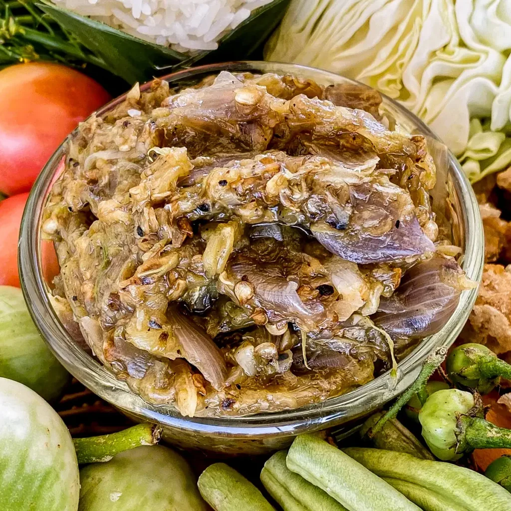Close-up of nam prik noom, a Northern Thai roasted chili dip, in a glass cup. Above and underneath the dip are parts of fresh vegetables and pork crackling.