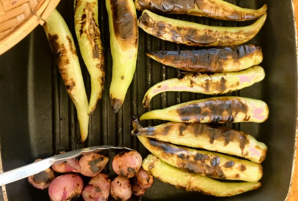 Top-down view of a grilling pan with roasted large green chilies and shallots.