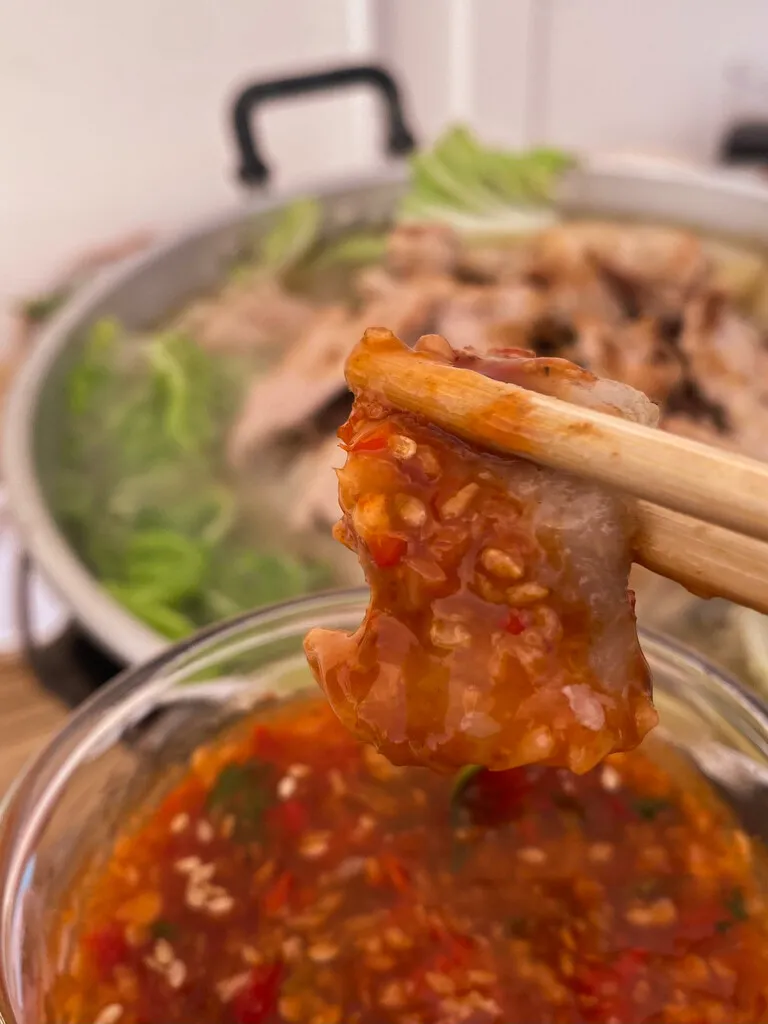 Holding a piece of thinly cut pork meat between chopsticks, dipped in a spicy Thai barbecue sauce, with a blurred Thai barbecue on the background.