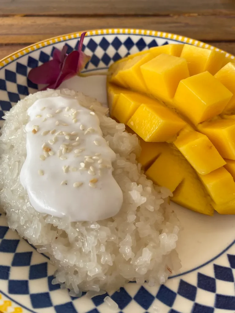 Mango with sticky rice, coconut milk topping sauce on top of the rice with toasted sesame seeds, and a purple flower in a white plate on a wooden background.