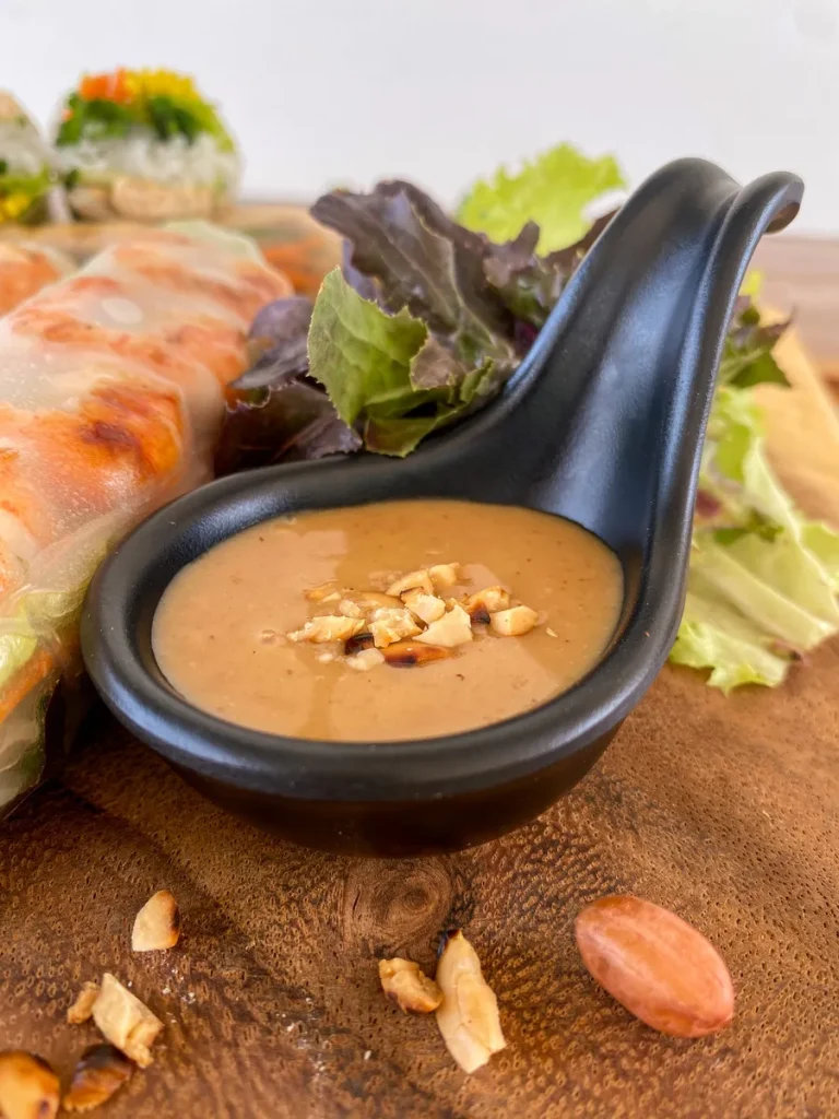 Peanut sauce in a black spoon with crumbs of peanut in the center. To the left there are fresh spring rolls, and behind it is lettuce.