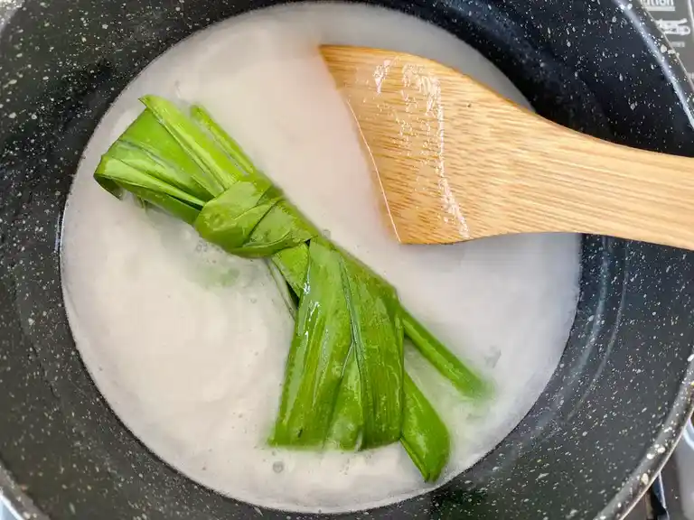 Coconut milk sauce being cooked with pandan leaves in a pot with a wooden spatula.
