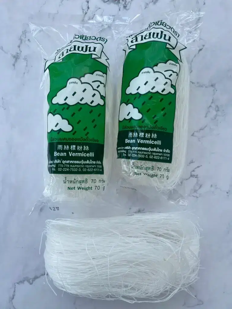 2 packages of glass noodles, and unpacked glass noodles underneath it, on a white background.