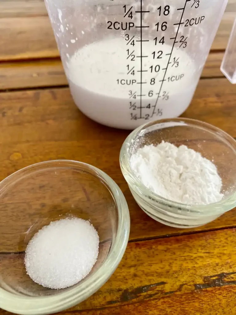 Measuring cup filled with coconut milk alongside small bowls of sugar and rice flour on a wooden table.