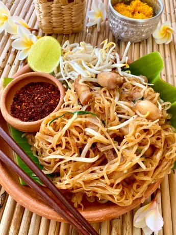 Pad mee korat on top of a banana leaf in a clay serving tray. On top of it is some dried chili flakes with wooden chopsticks, a wedge of lime, and flowers are spread around as decoration.