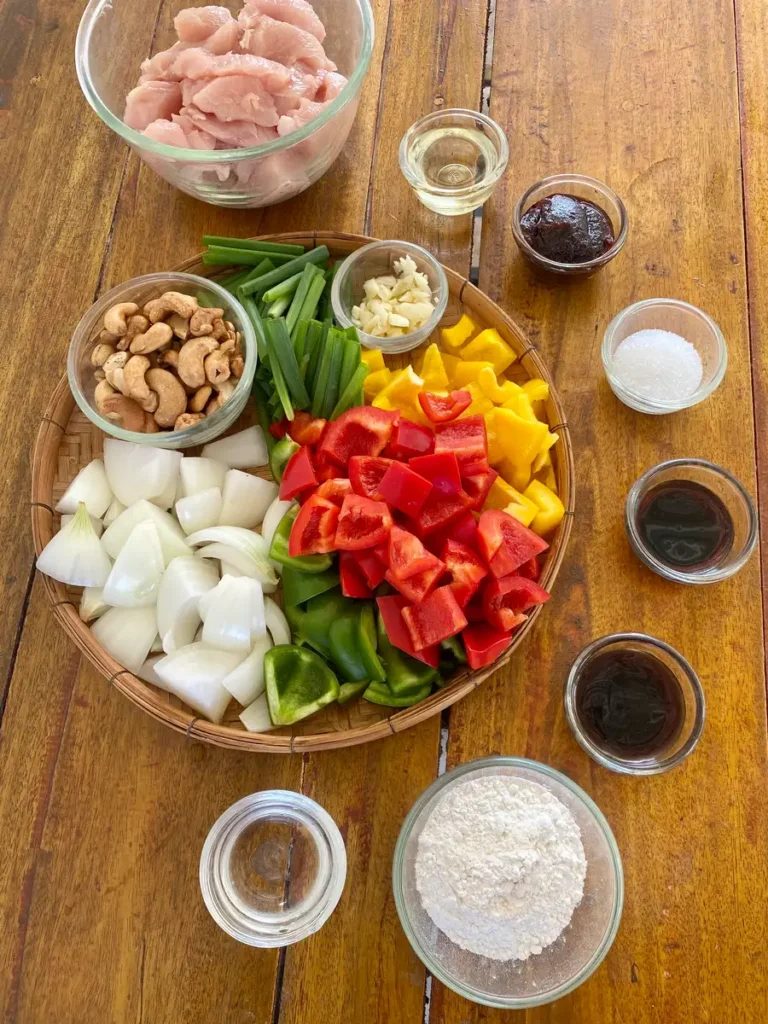 Bird's eye view of recipe ingredients; crispy flour, water, oyster sauce, soy sauce, sugar, vegetables, chicken, chili paste, oil, and cashews.