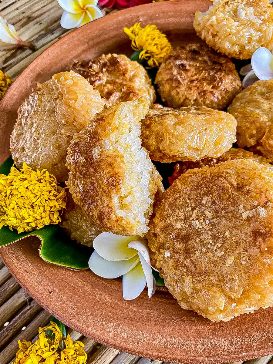 Crispy grilled sticky rice patties coated in egg, served on a clay plate with banana leaf and fresh flowers.