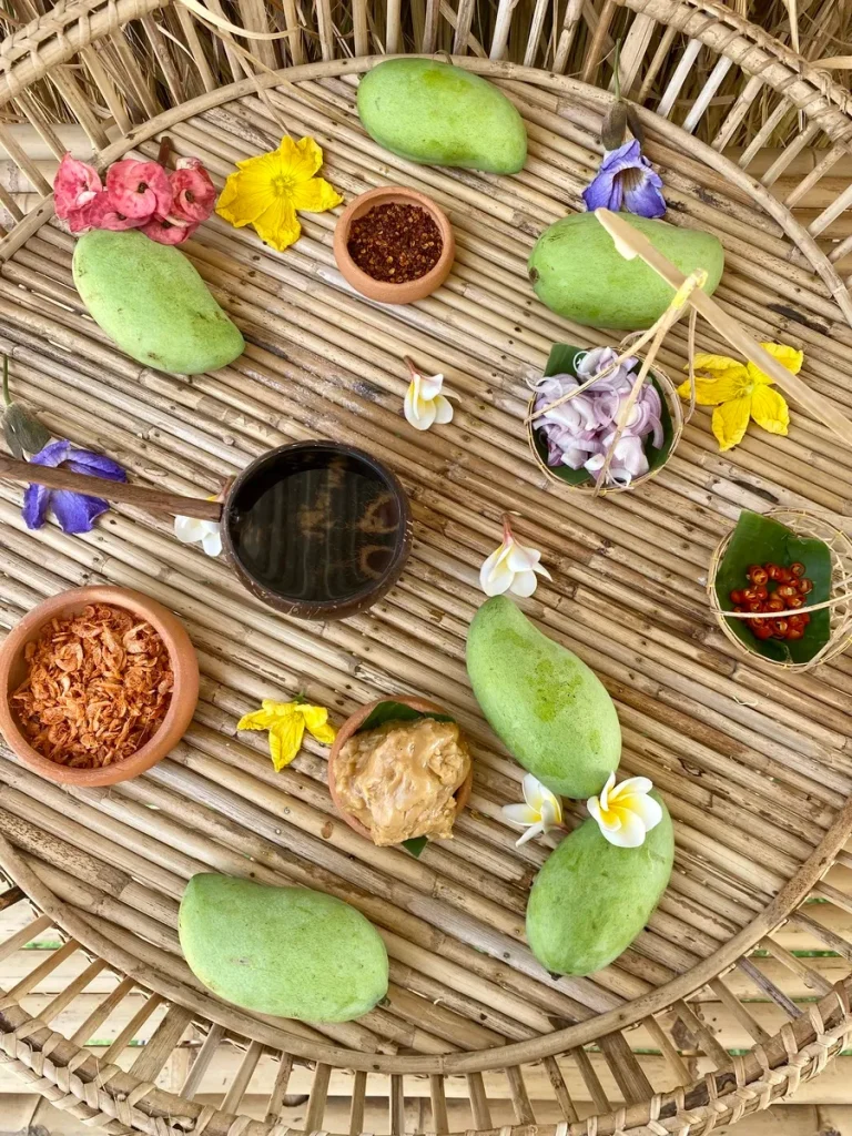 Ingredients for mango dipping sauce: Green mangoes, dried chili flakes, flowers, shallots, chopped red chili, a wooden spoon with water, dried shrimp, and palm sugar on top of a bamboo serving tray.
