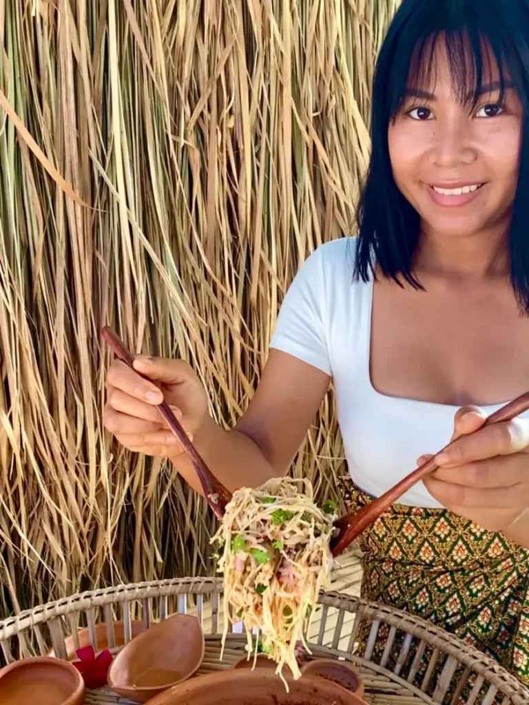 Smiling Thai woman enjoying a fresh enoki mushroom larb salad, using wooden chopsticks, with a backdrop of dried grasses.