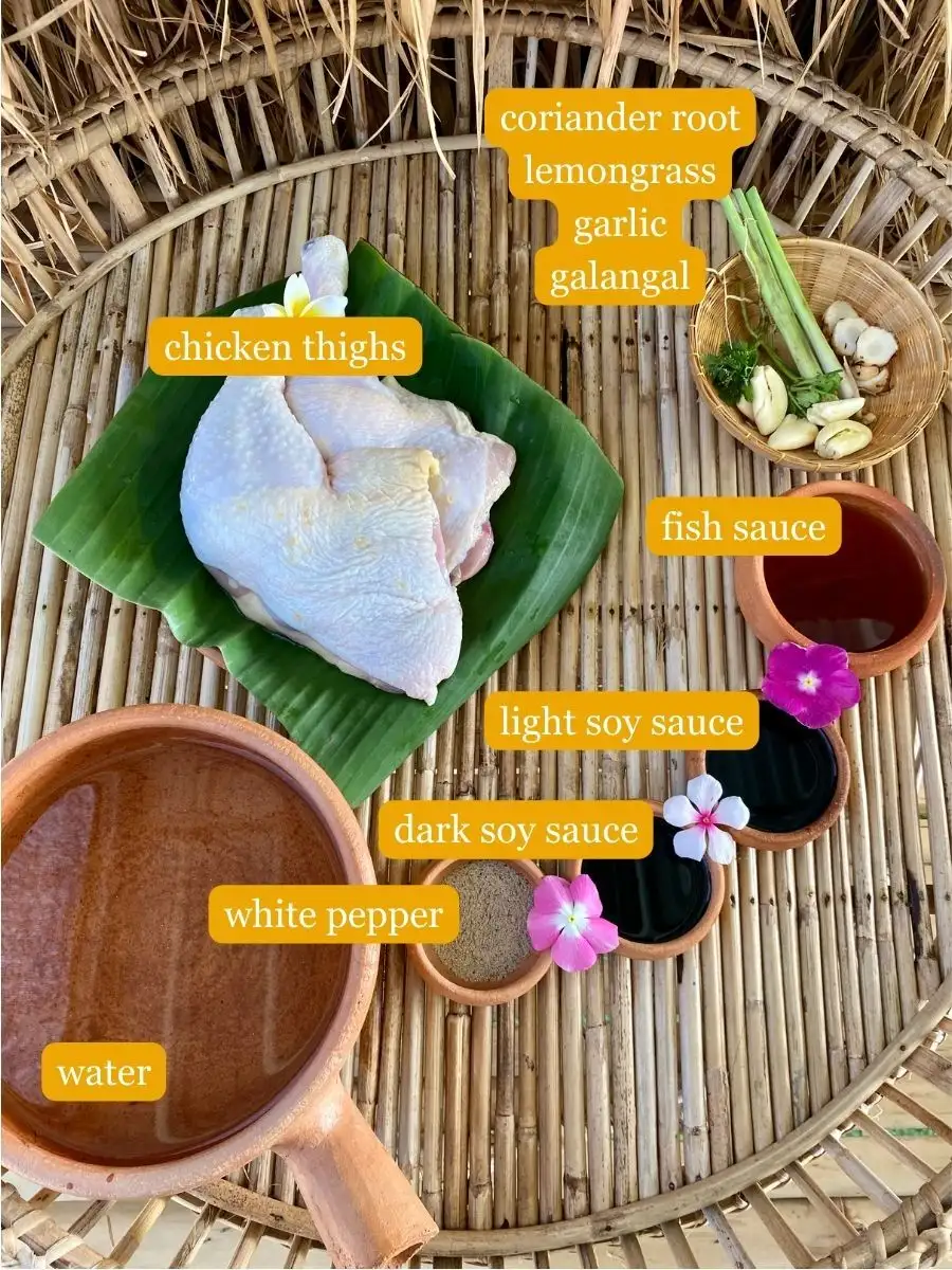 All ingredients for Thai boiled chicken on a bamboo serving tray; raw chicken thighs, fish sauce, dark soy sauce, light soy sauce, palm sugar, galangal, lemongrass, garlic, coriander root, water, and white pepper.