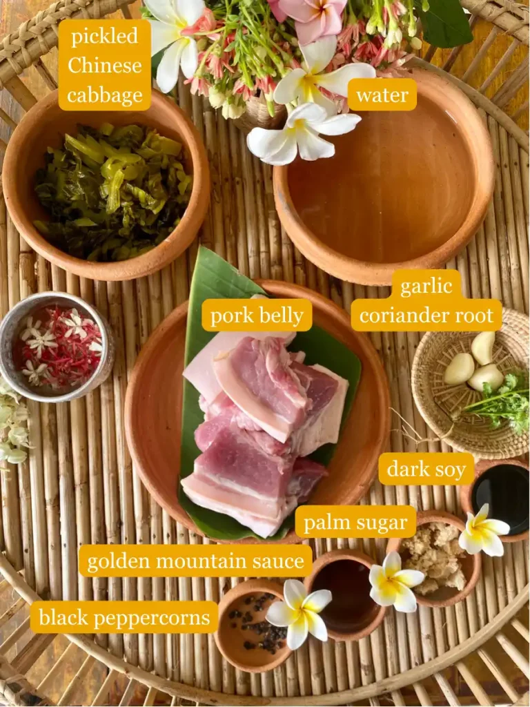 Top-view of recipe ingredients: Pickled Chinese cabbage, water, raw pork belly, garlic, coriander root, dark soy sauce, palm sugar, golden mountain sauce, black peppercorns on a bamboo serving tray.