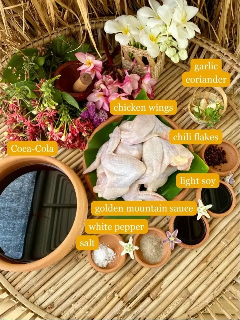 Salt, white pepper, golden mountain sauce, light soy sauce, chili flakes, raw chicken wings, coca-cola, garlic, and coriander on a bamboo serving tray with flowers.