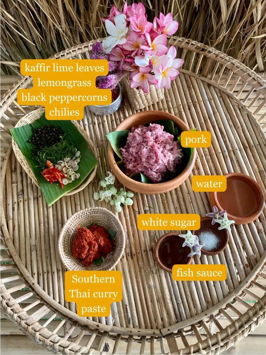 An overhead shot of all ingredients for khua kling on a bamboo serving tray; kaffir lime leaves, lemongrass, black peppercorns, chilies, minced pork, water, white sugar, water, fish sauce, Southern Thai curry paste.