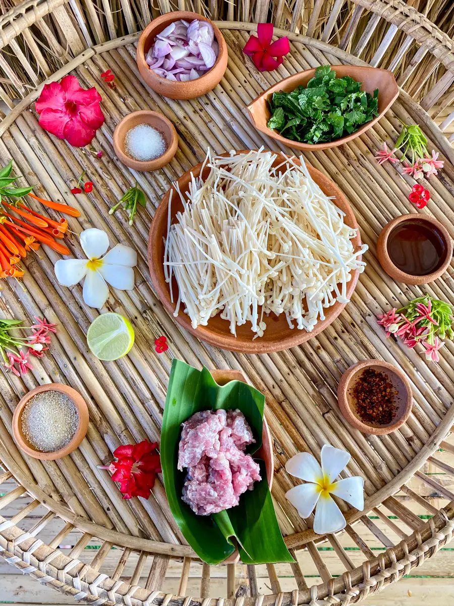 Ingredients for Thai mushroom salad (larb hed), including enoki mushrooms, ground pork, herbs, shallots, fish sauce, lime, and toasted rice powder.