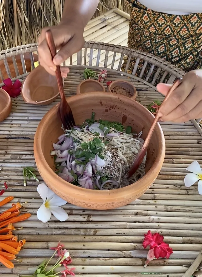 Mixing enoki mushroom larb salad with both my hands and a wooden fork and spoon, in a clay bowl on top of a bamboo serving tray that has flowers in it.