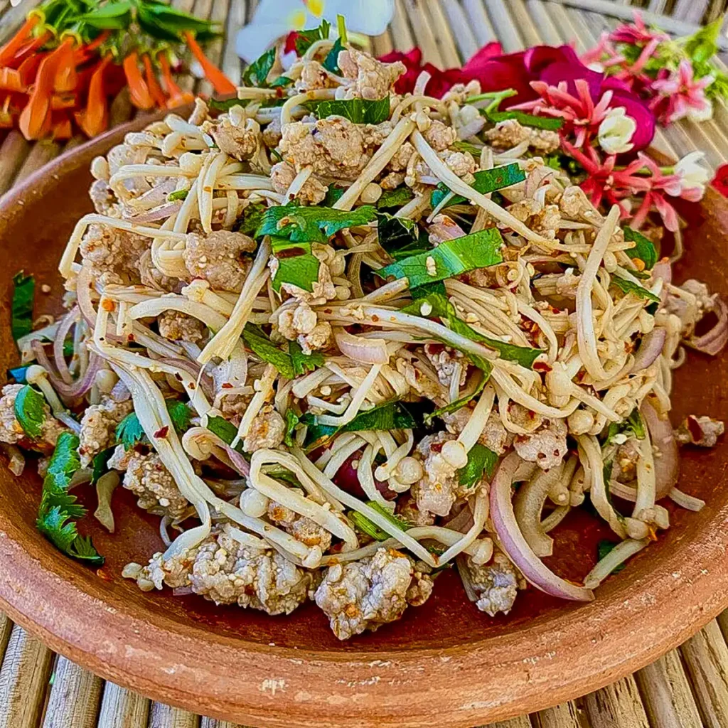 Close-up of spicy mushroom larb with minced meat, herbs, enoki mushrooms, and toasted rice powder served in a rustic clay plate.