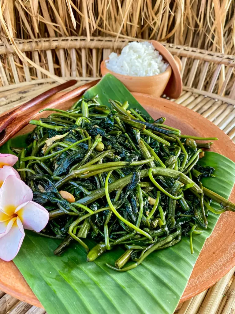 Stir-fried Thai morning glory (pad pak boong) on a clay plate lined with banana leaf, served with rice in a clay bowl.