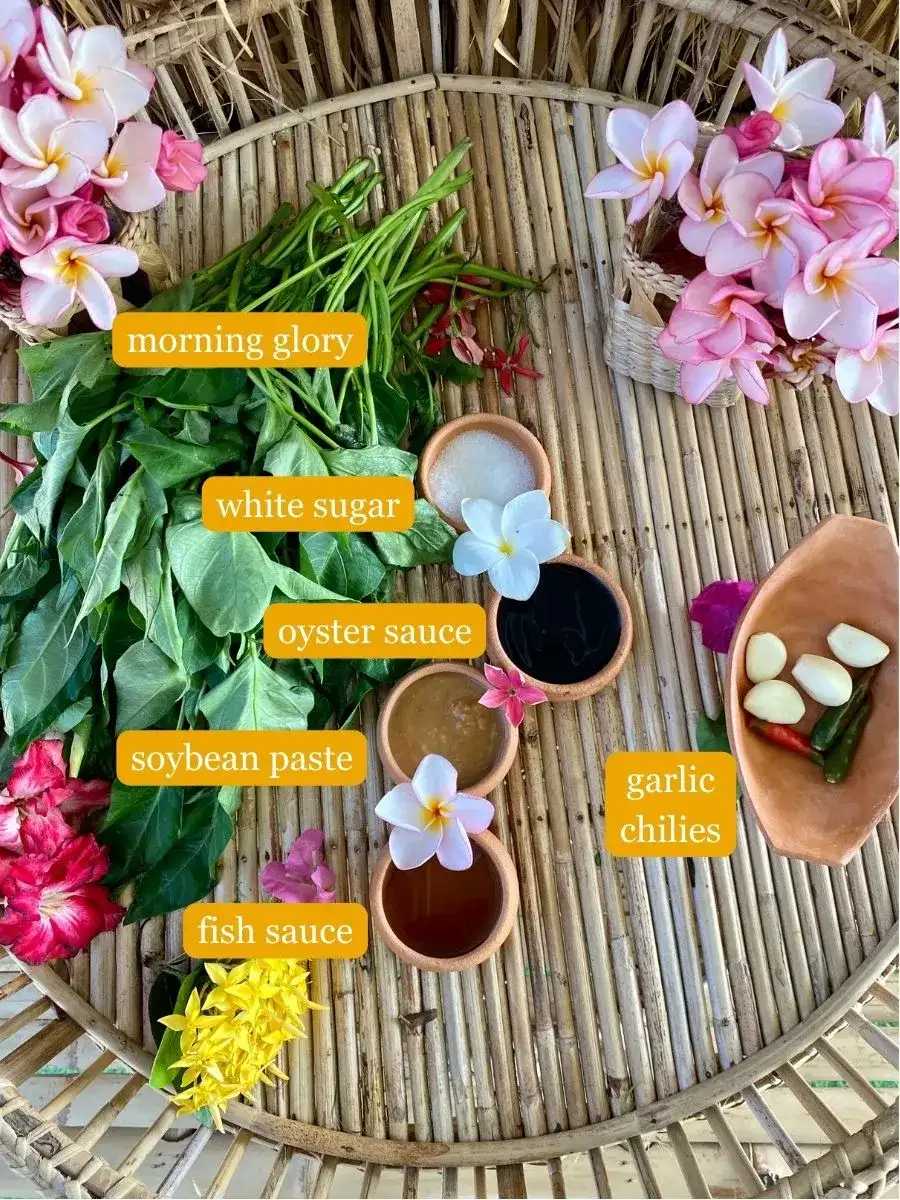 Overhead view of ingredients on a bamboo tray. Crispy morning glory, white sugar, oyster sauce, soybean paste, fish sauce, and a garlic chili combo.