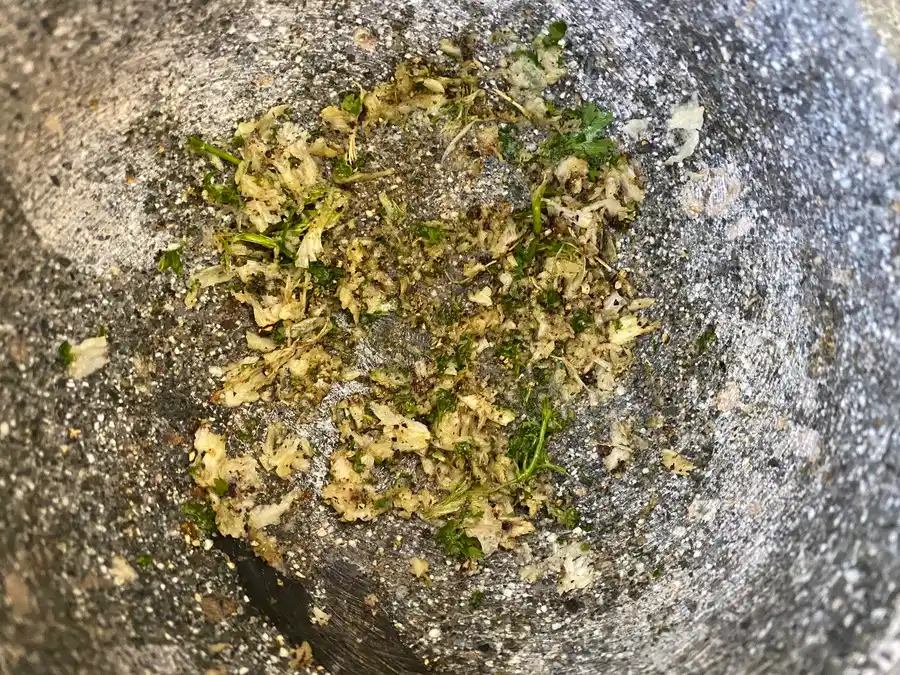 Close-up of pounded garlic, coriander root, and black peppercorns in a granite mortar.
