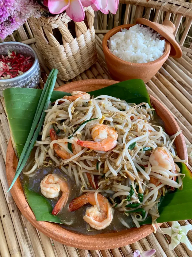 Shrimp with bean sprouts stir-fry on a banana leaf in a clay dish. Above it is a portion of white rice and flowers as decoration.