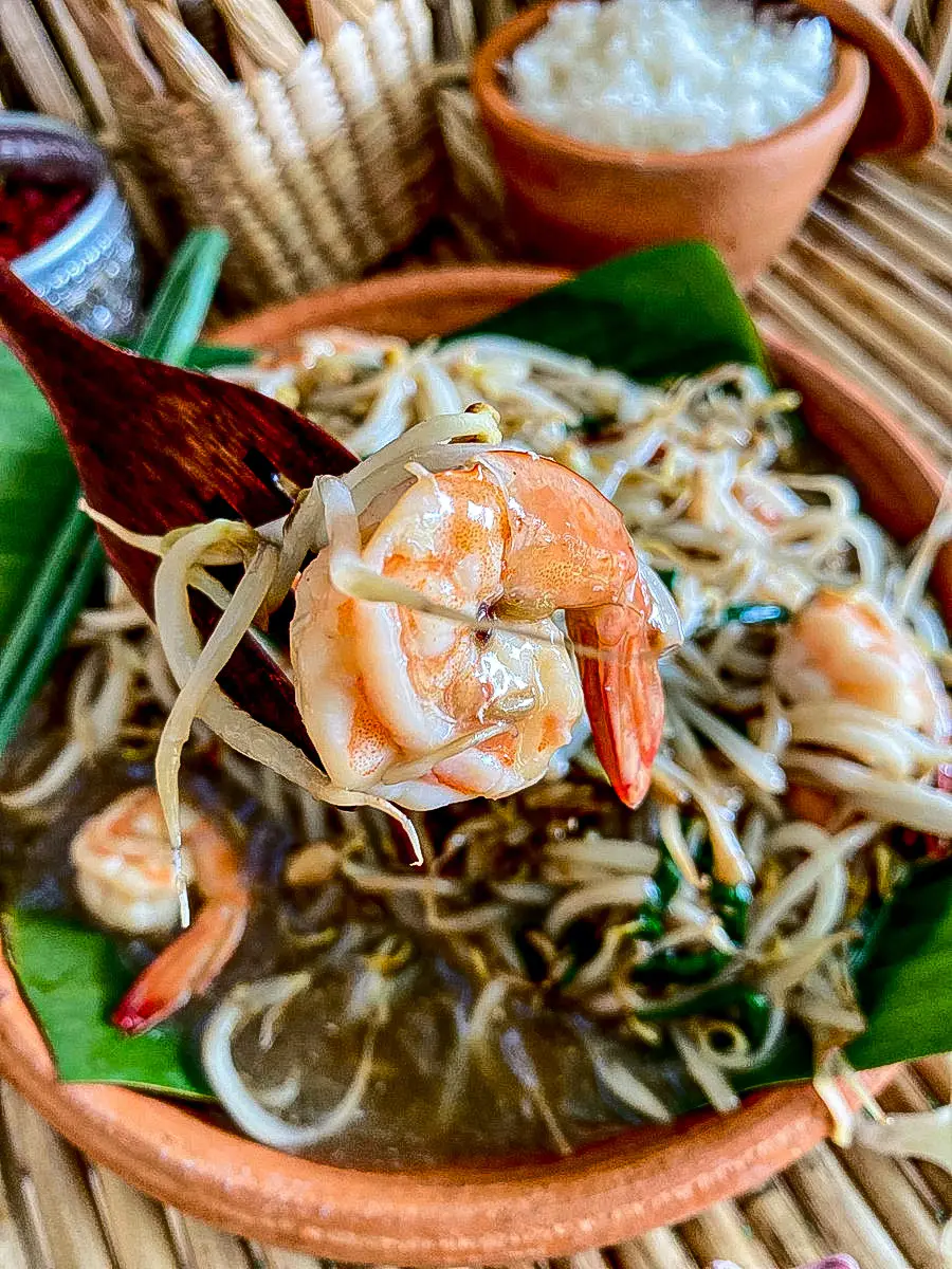 Wooden fork holding shrimp over clay plate with bean sprouts stir fry.