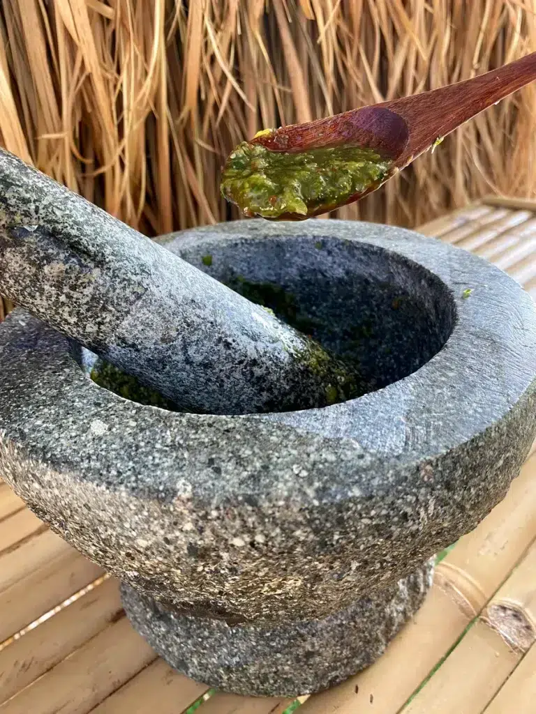Green nam jim seafood dipping sauce in a stone mortar and pestle on top of a bamboo table, above it is a wooden spoon that holds some sauce in it.