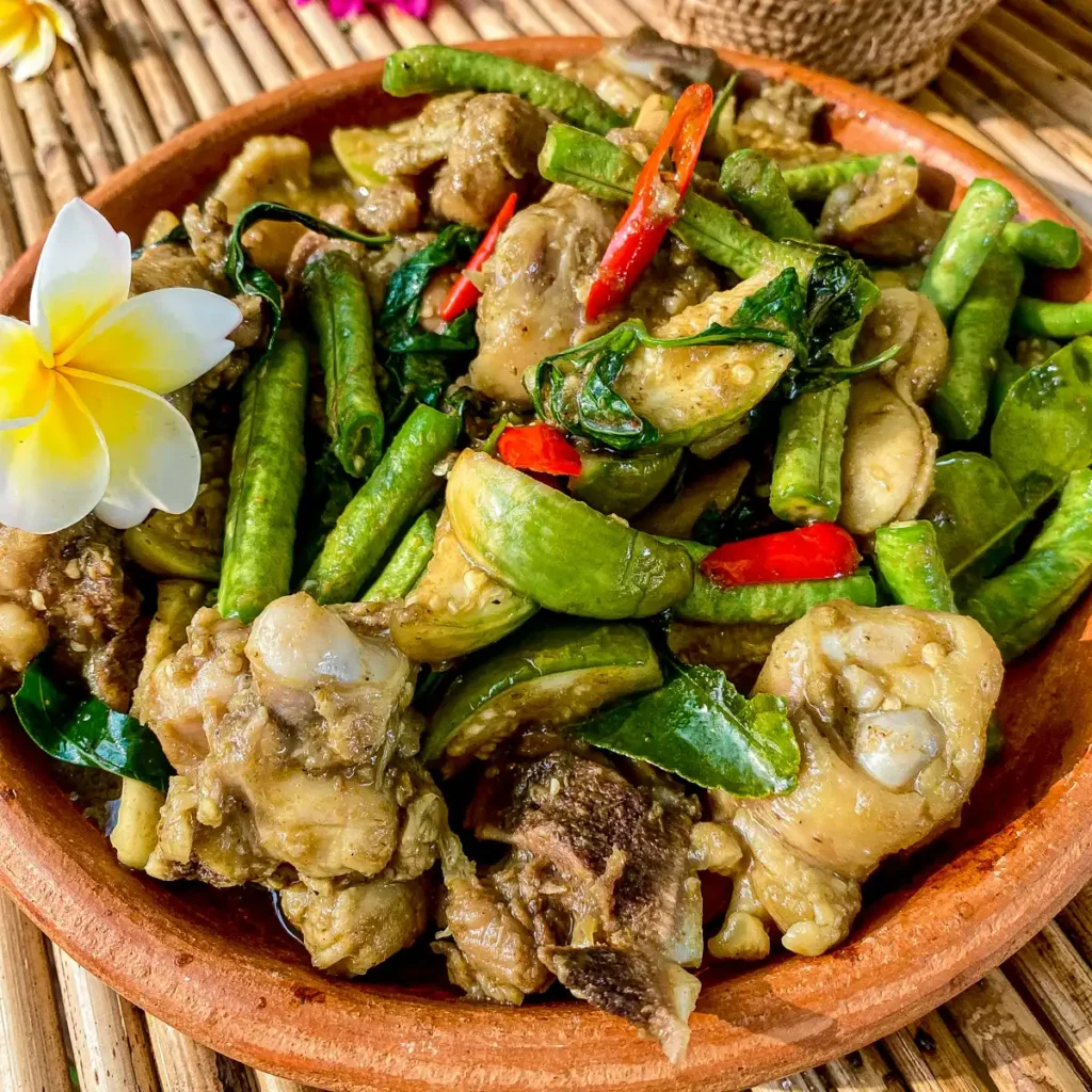 Thai green curry stir fry with chicken, Thai eggplants, long beans, and red chilies served in a clay bowl with tropical flowers.