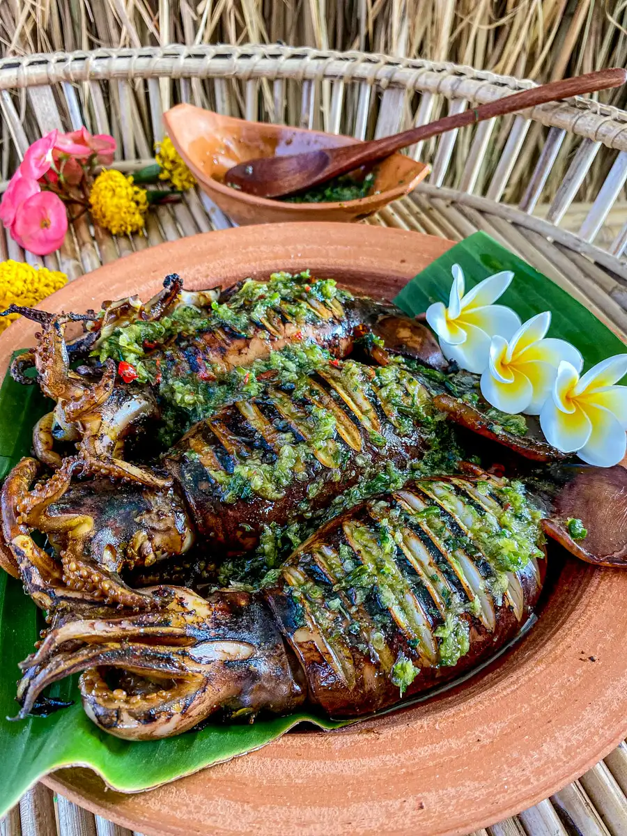 Thai grilled squid served on a banana leaf, drizzled with vibrant green seafood sauce, garnished with plumeria flowers, and set in a rustic clay plate.