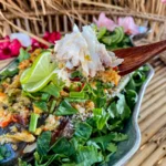Thai lime garlic steamed fish topped with fresh coriander, Chinese celery, chilies, and lime wedges, with a serving of tender fish on a wooden spoon.