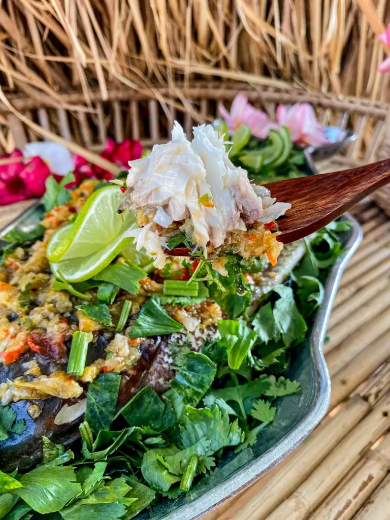 Thai lime garlic steamed fish topped with fresh coriander, Chinese celery, chilies, and lime wedges, with a serving of tender fish on a wooden spoon.