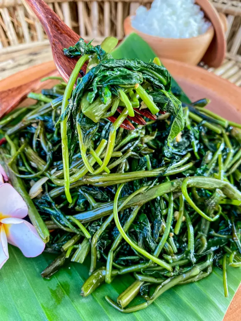 A fork lifting stir-fried Thai morning glory with garlic and soy sauce, served on a banana leaf with jasmine rice in the background.