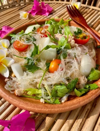 Yum woon sen pork (Thai glass noodle salad) in a clay bowl with a wooden fork on top, surrounded by flowers, on a bamboo serving tray.