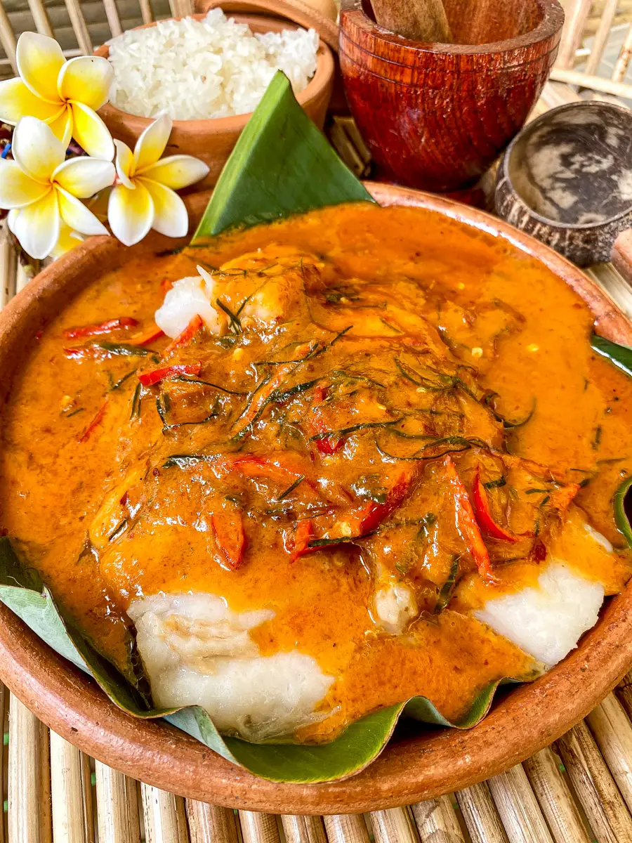 Thai red curry fish served in a clay bowl with banana leaf lining, topped with julienned herbs and sliced red chili.