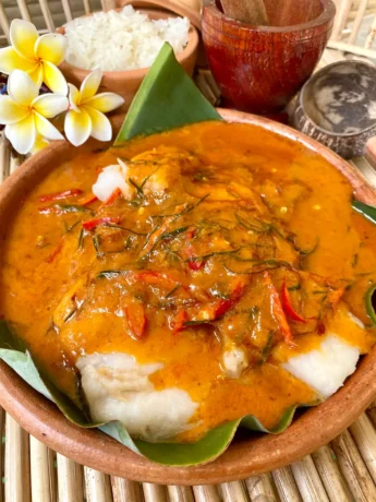 Thai red curry fish in a clay dish with red chilies and strips of kaffir lime leaves. There's also a portion of white rice, flowers, and a small mortar and pestle.