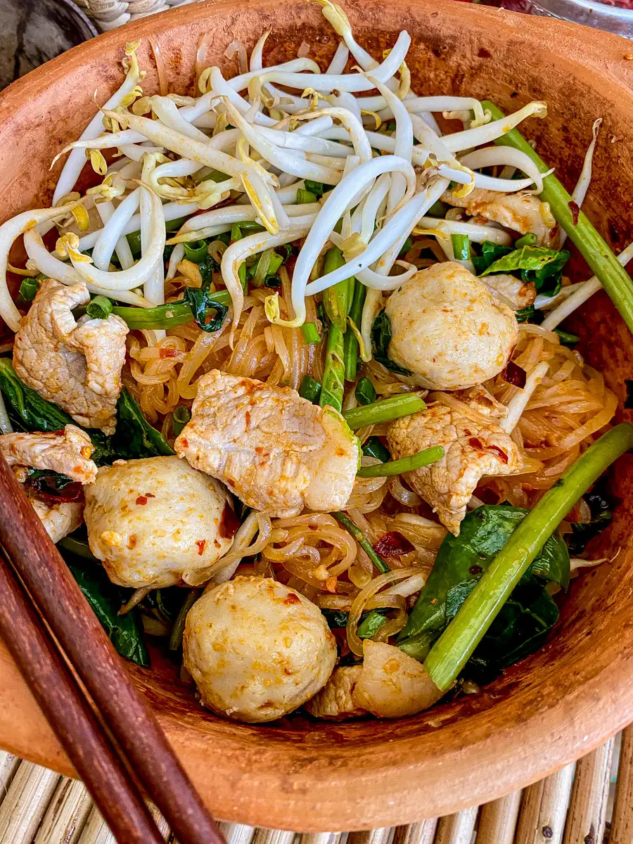 Close-up of spicy dry tom yum noodles with pork, fish balls, morning glory, and bean sprouts in a clay bowl.