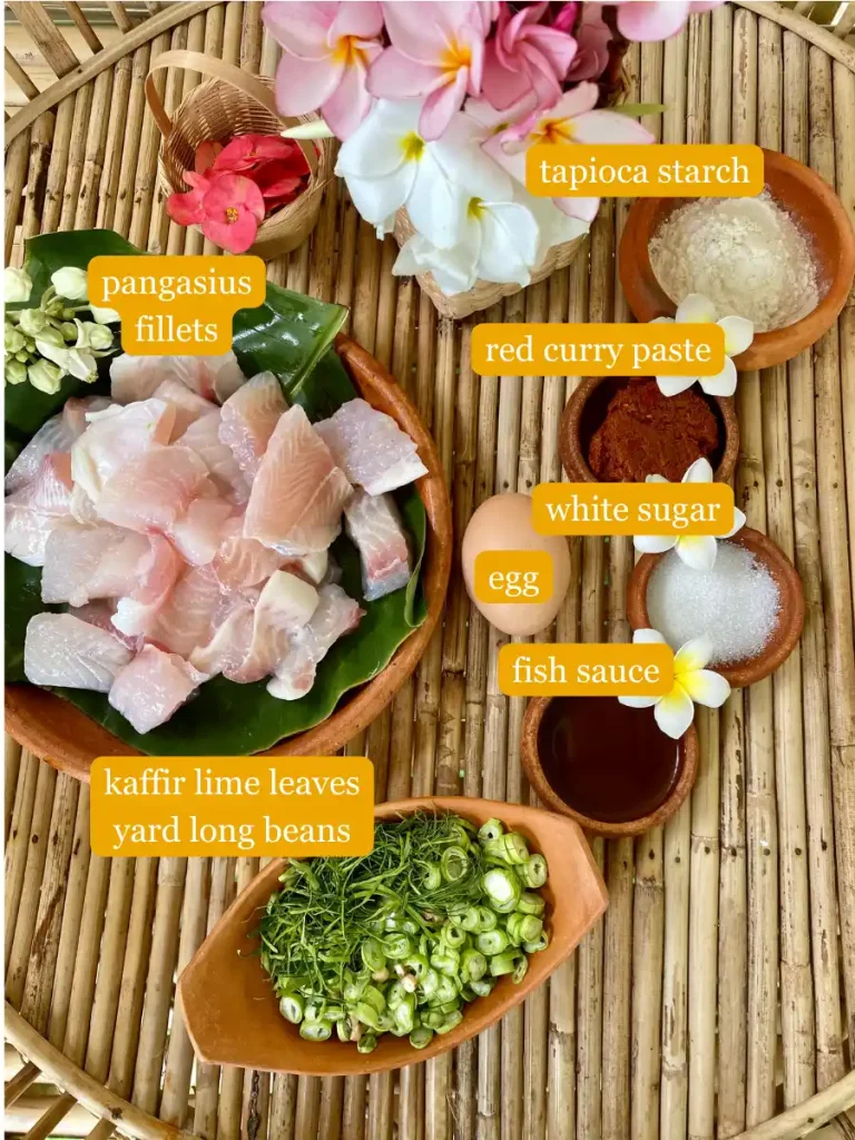 Top view of tod mun pla ingredients: Tapioca starch, pangasius fillets, red curry paste, white sugar, an egg, fish sauce, sliced kaffir lime leaves, sliced yard long beans, and flowers on a bamboo serving tray.